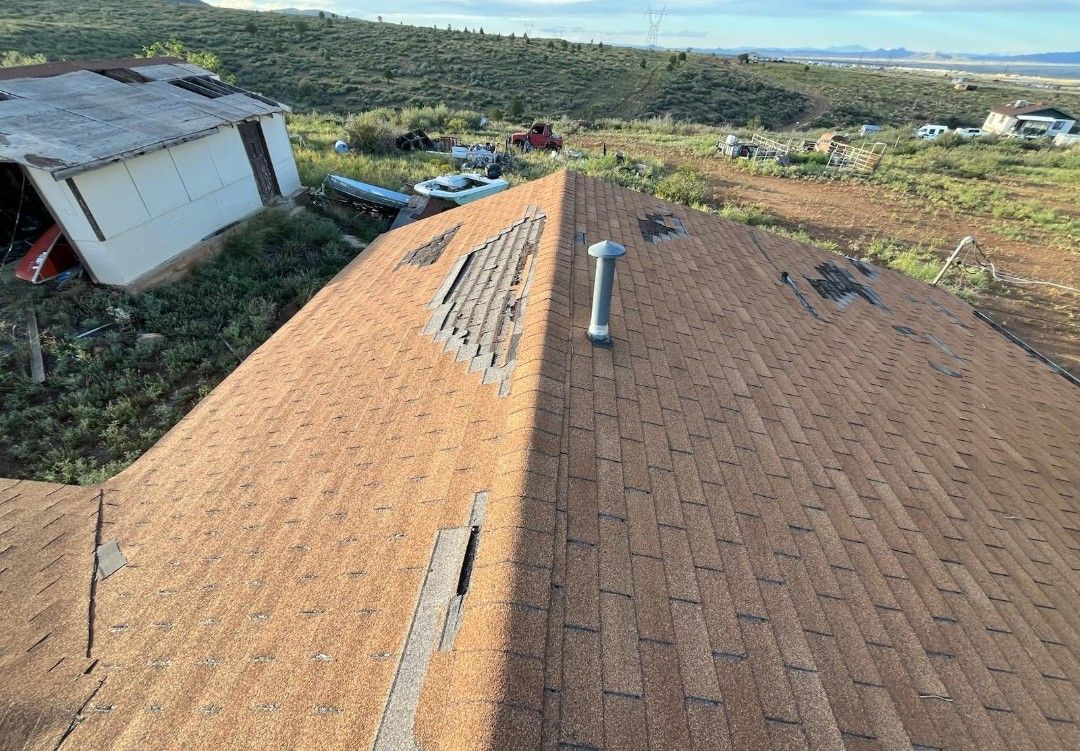 Damaged brown shingle roof with missing sections, view from above. Adjacent to a white shed and grassy field.