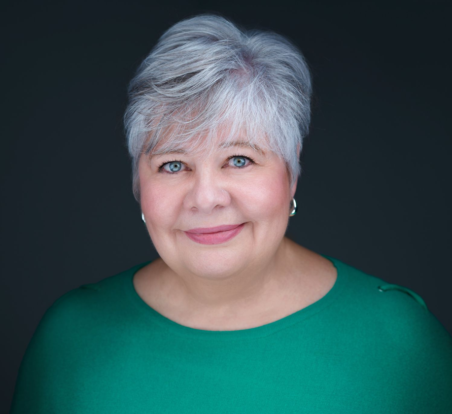 Studio headshot of a smiling person with short silver hair against a dark background