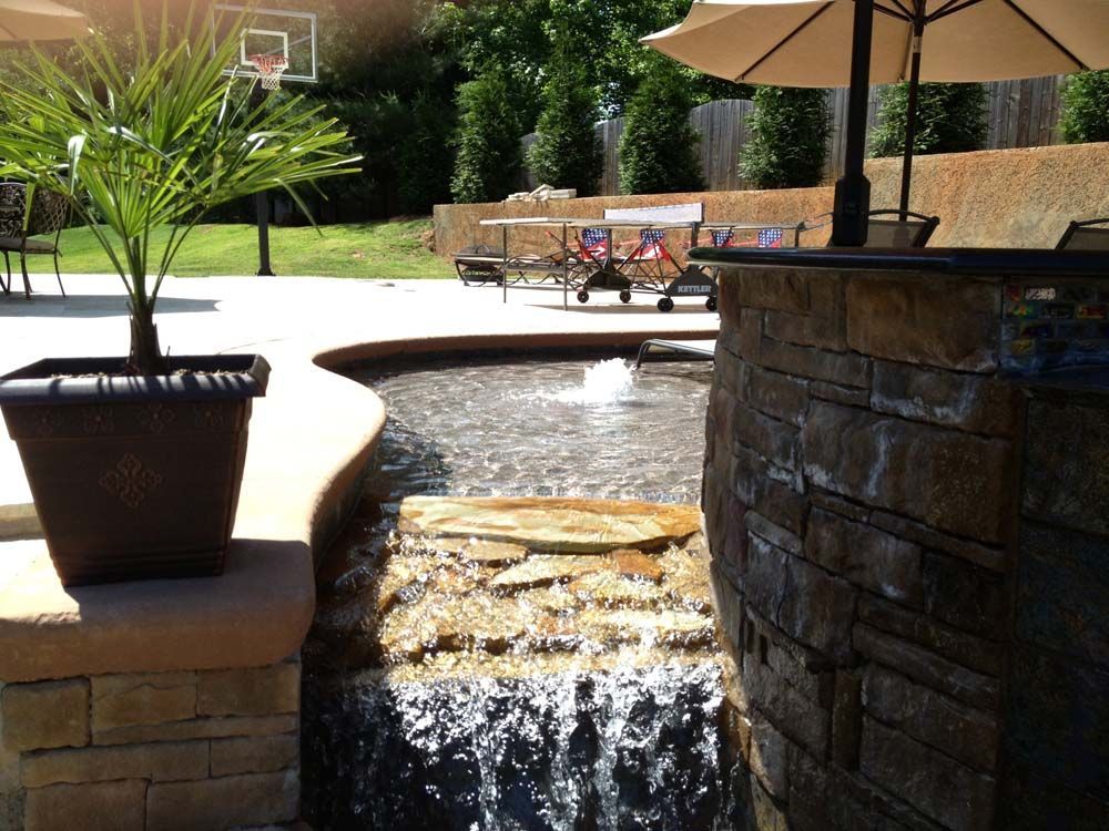Water feature with waterfall flowing into a pool, bordered by a stone wall and potted palm, outdoor setting.