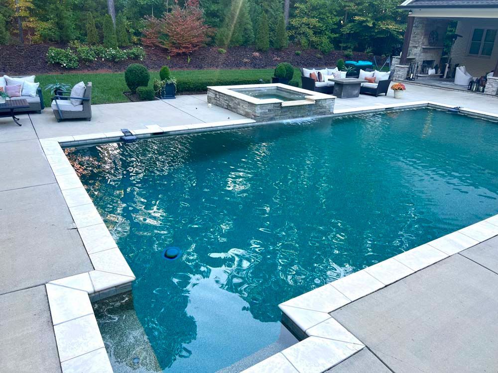 Backyard swimming pool with stone coping, lounge chairs, and a raised spa near lush greenery