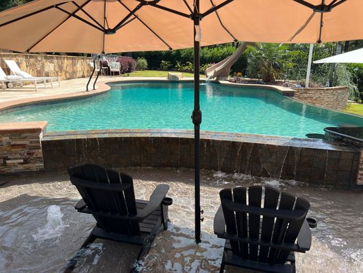Two chairs under an umbrella facing a turquoise swimming pool with a stone waterfall edge