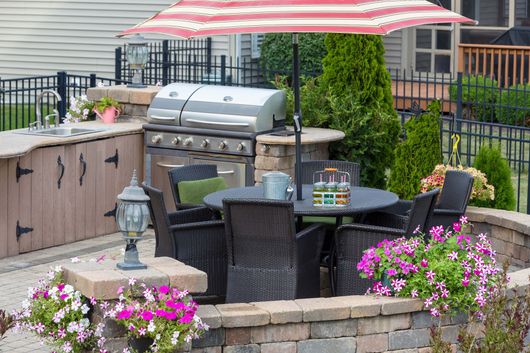 Backyard patio with grill, table and chairs under a striped umbrella, surrounded by flowers and a stone wall