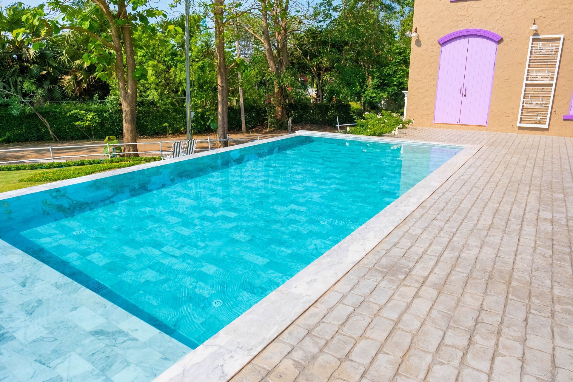Rectangular turquoise swimming pool beside a brick patio and trees, with a purple door on the building wall