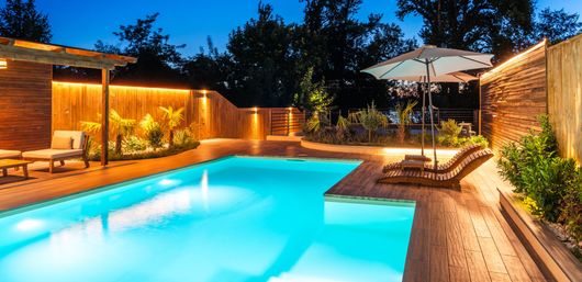 Glowing backyard pool at dusk with loungers, umbrellas, and warm wooden decking
