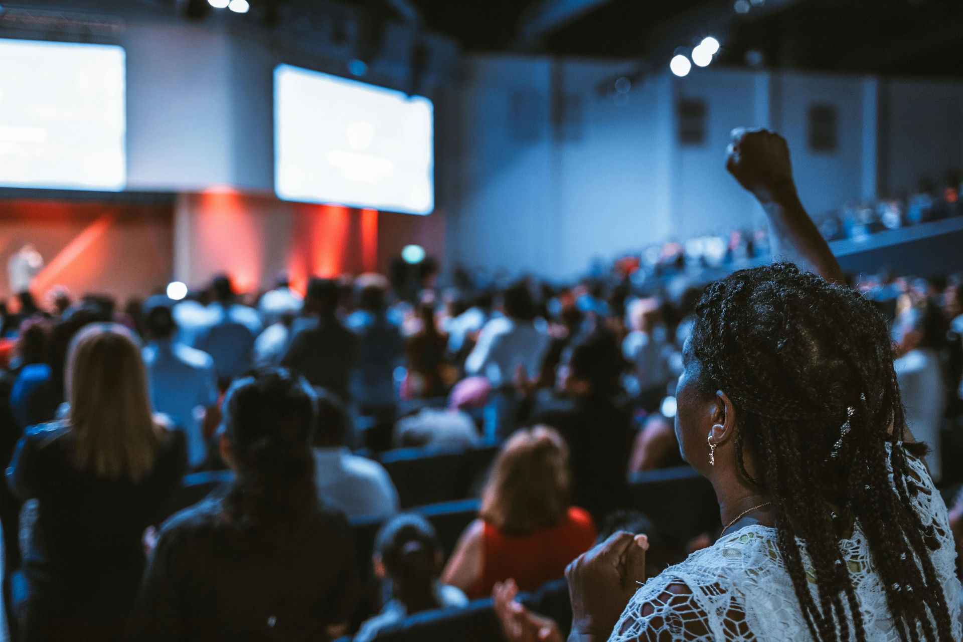 Woman with fist raised in a large crowded auditorium, focused on stage with screens.