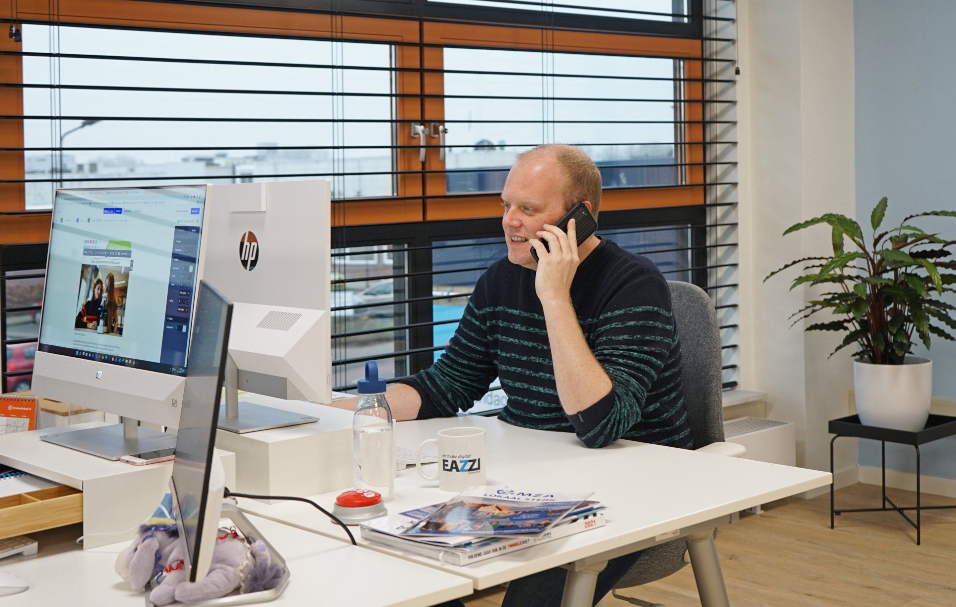 Arno zit aan een bureau en praat op een mobiele telefoon.