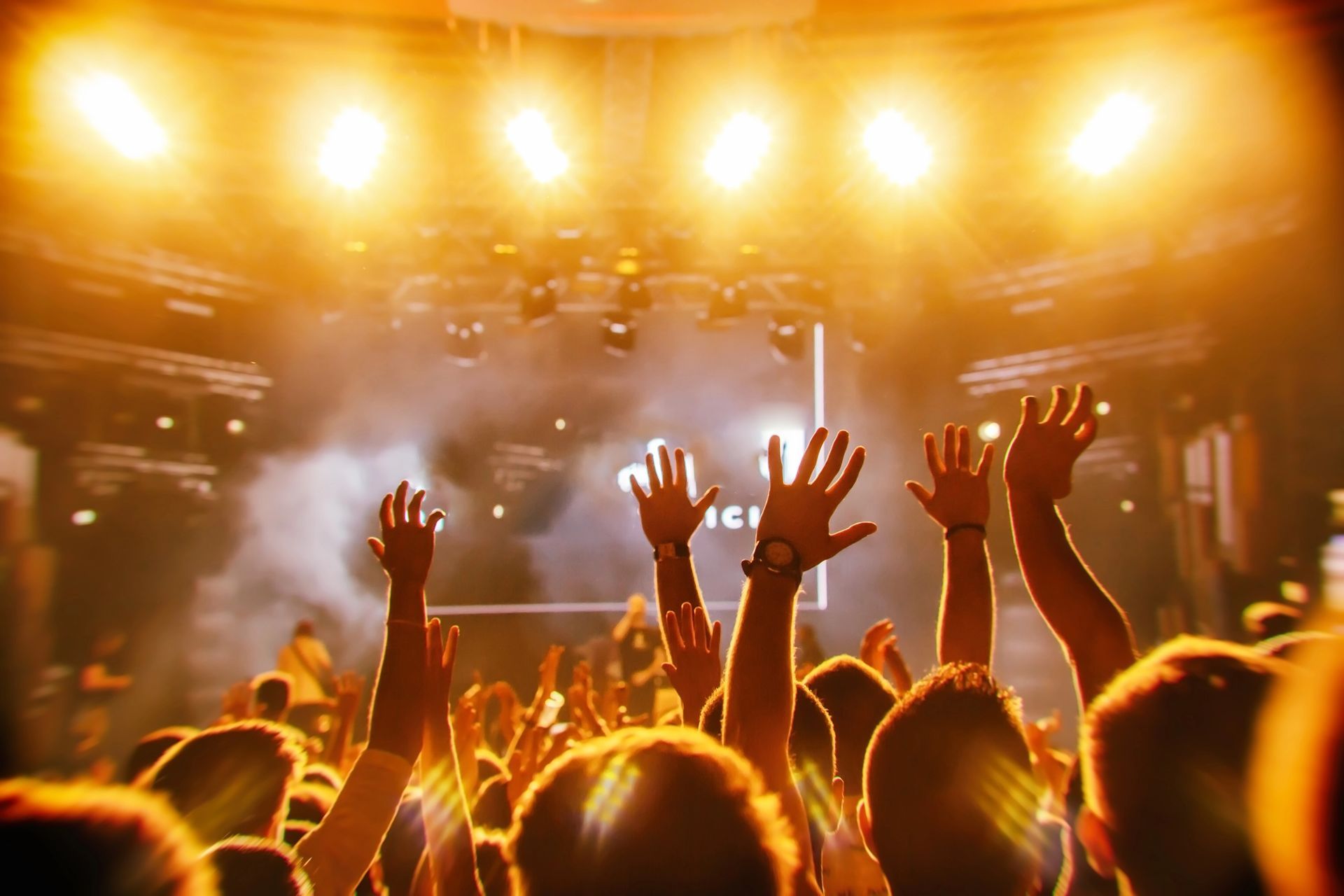 Pubblico con le mani alzate durante un concerto sotto le brillanti luci dorate del palco.