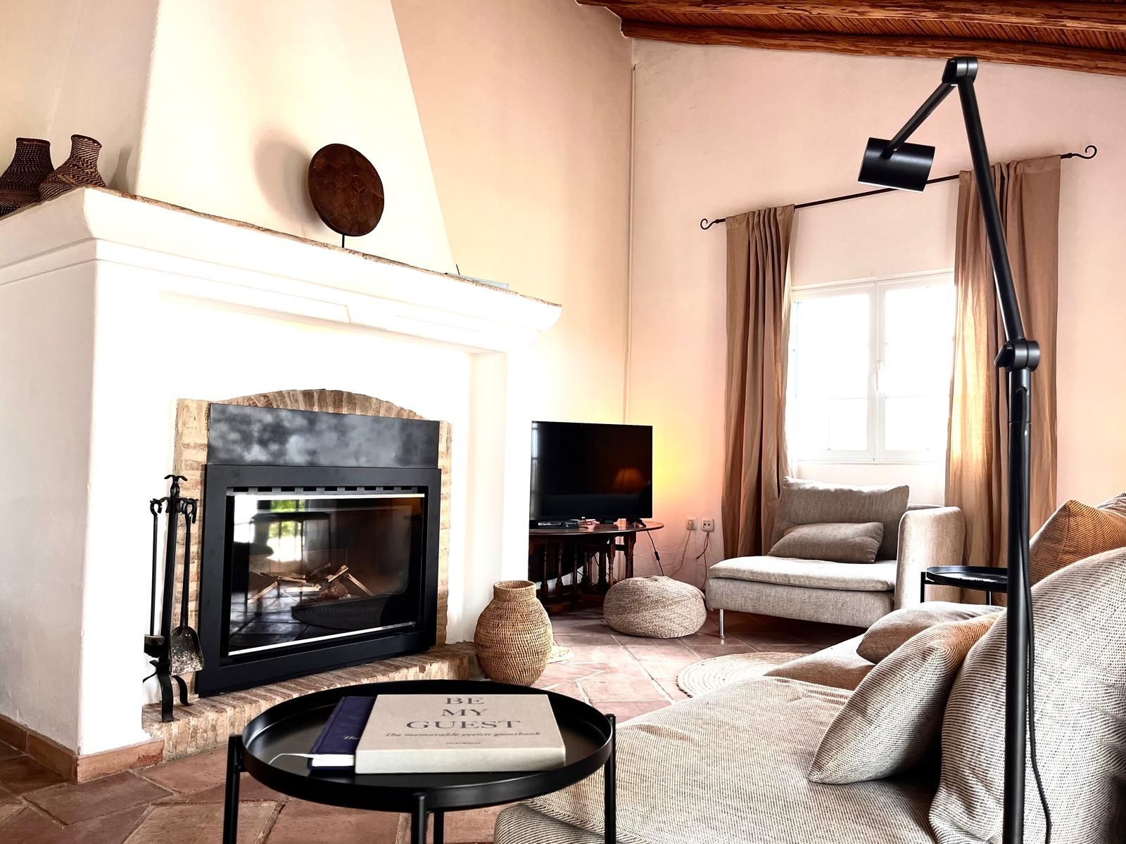 Living room with fireplace, couch, armchair, and black lamp. Beige walls, brown curtains, and wood ceiling.