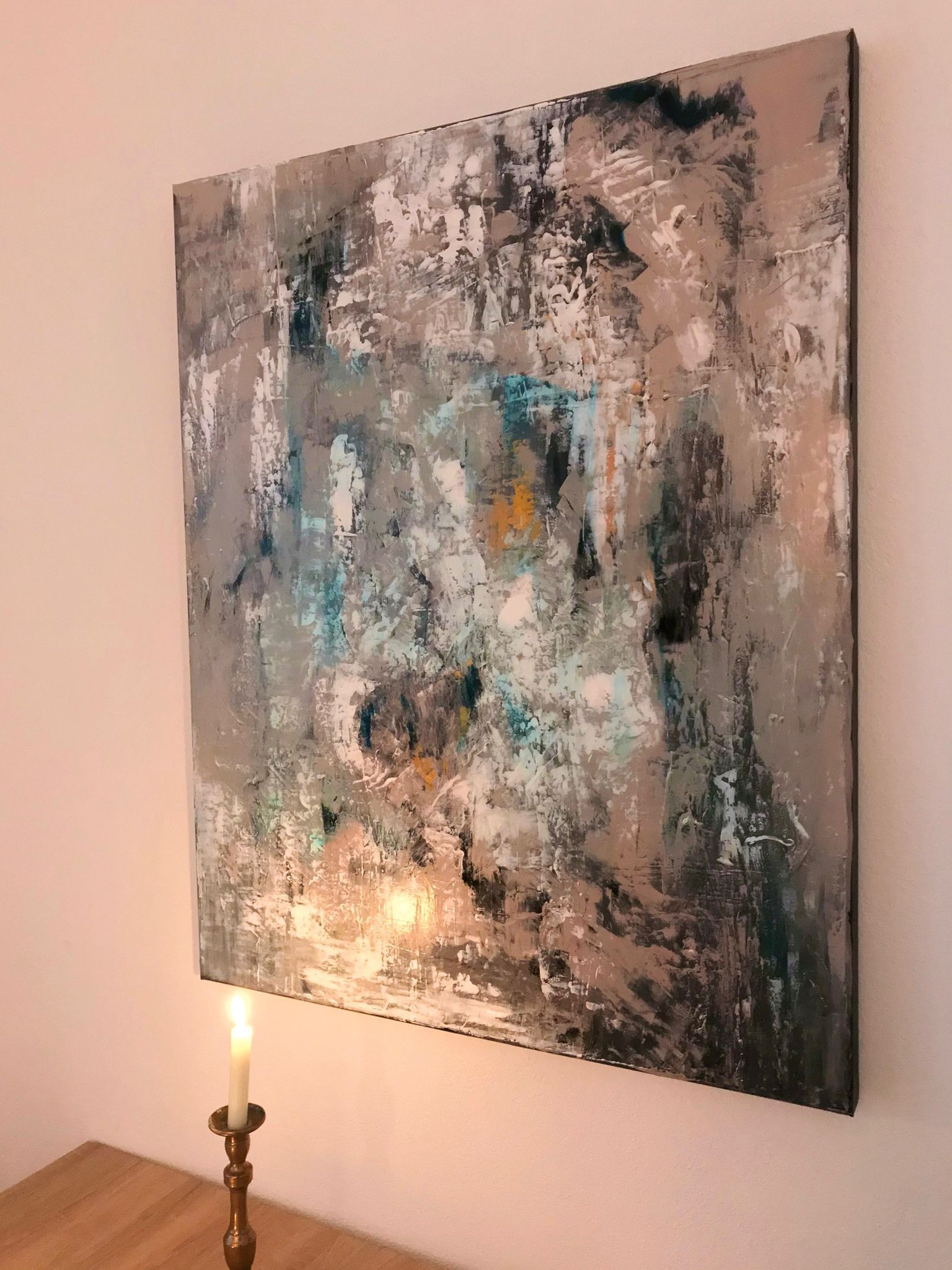 Abstract painting on a white wall, with teal, grey, and white brushstrokes, lit by a candle.
