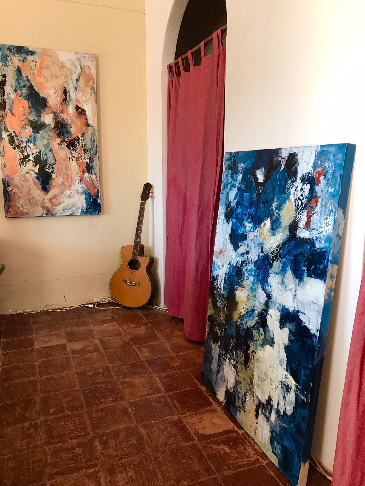 A room with art. A guitar leans on the wall. Paintings in blue, white, and peach tones. Red curtains in the doorway.
