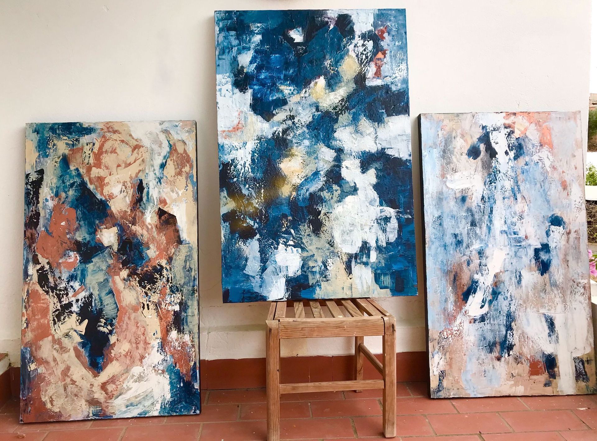 Three abstract paintings in blue, brown, and white lean against a wall. One stands on a wooden stool.