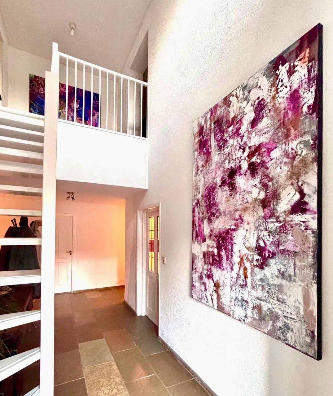 An abstract painting in shades of pink, white and grey hanging on a white wall in a bright hallway with a staircase.