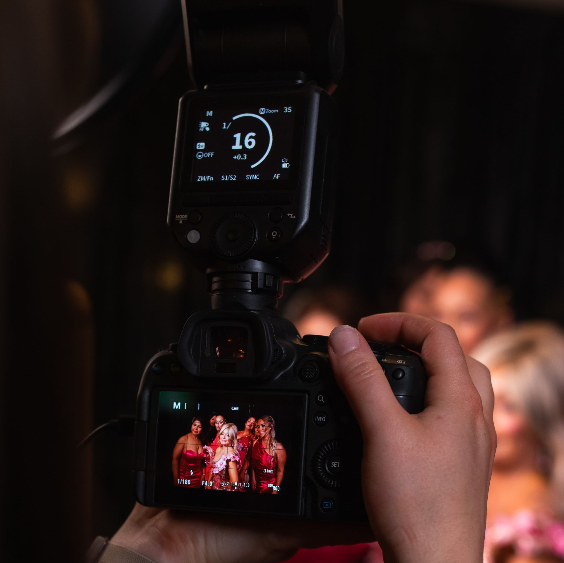 Photographer taking a photo of people; camera flash, screen showing subjects in red. Dark setting. Event Photography.