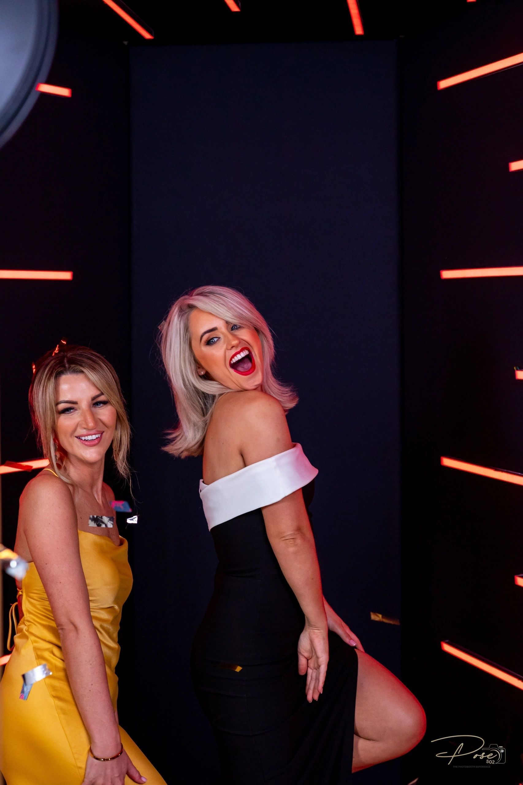 Two women in dresses are posing for a picture in a photo booth.
