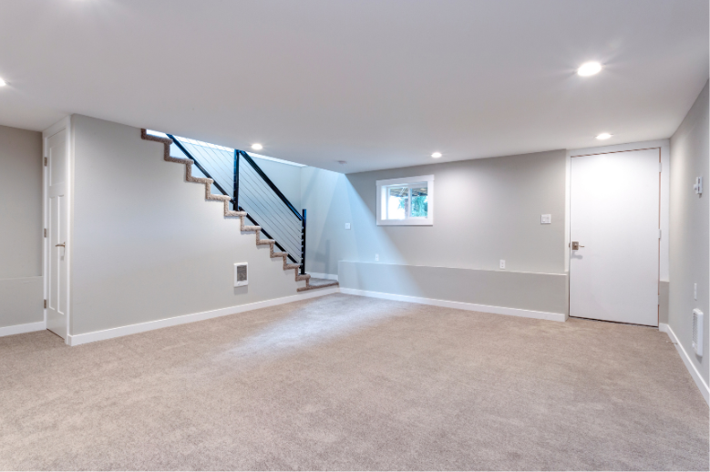 Empty carpeted basement room with gray walls, a staircase, small window, and white door