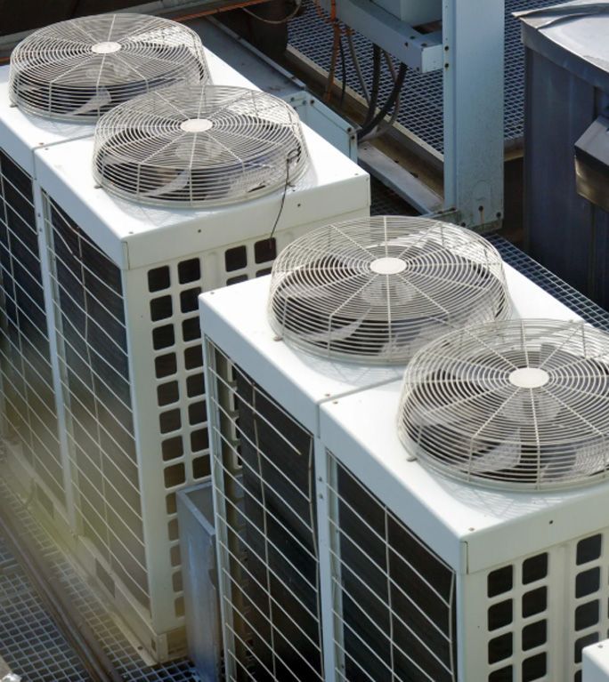Four white, industrial outdoor air conditioning units with circular fan grilles, positioned on a rooftop.
