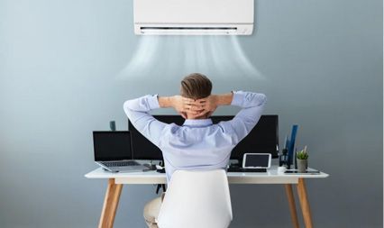 A person sits at a desk with hands behind their head, relaxing in front of a wall-mounted air conditioner.