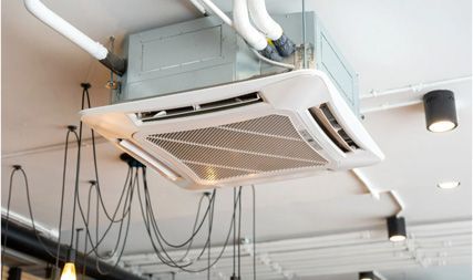 A white ceiling-mounted cassette air conditioning unit installed in a commercial space with exposed conduit and lighting.