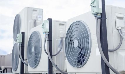 Several white outdoor air conditioning units arranged in a row, with connected electrical boxes and conduits.
