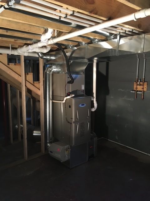 An HVAC furnace unit installed in a basement, featuring metal ductwork and white PVC pipes against a dark wall.