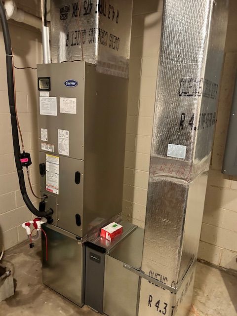 A gray indoor HVAC unit with silver ductwork stands in a basement against a concrete block wall.