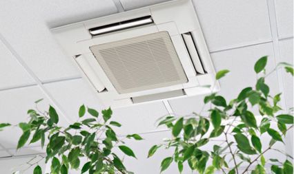 A white four-way ceiling cassette air conditioning unit mounted in a tiled office ceiling above green plants.