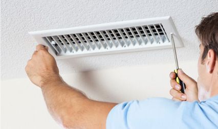 A person in a light blue shirt uses a screwdriver to install or repair a white rectangular ceiling air vent.