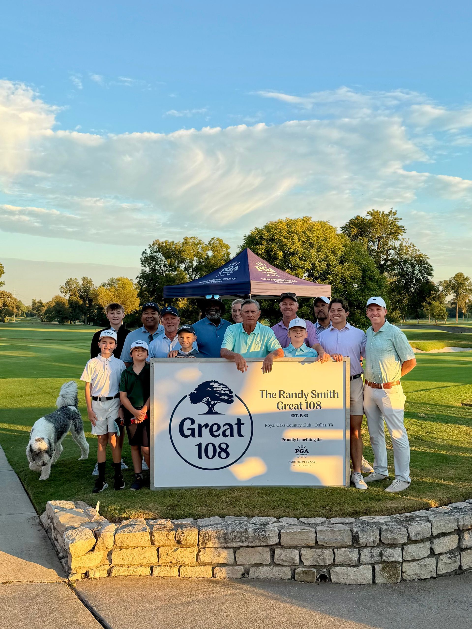Group posing beside Great 108 sign at an outdoor event under a blue tent at sunset