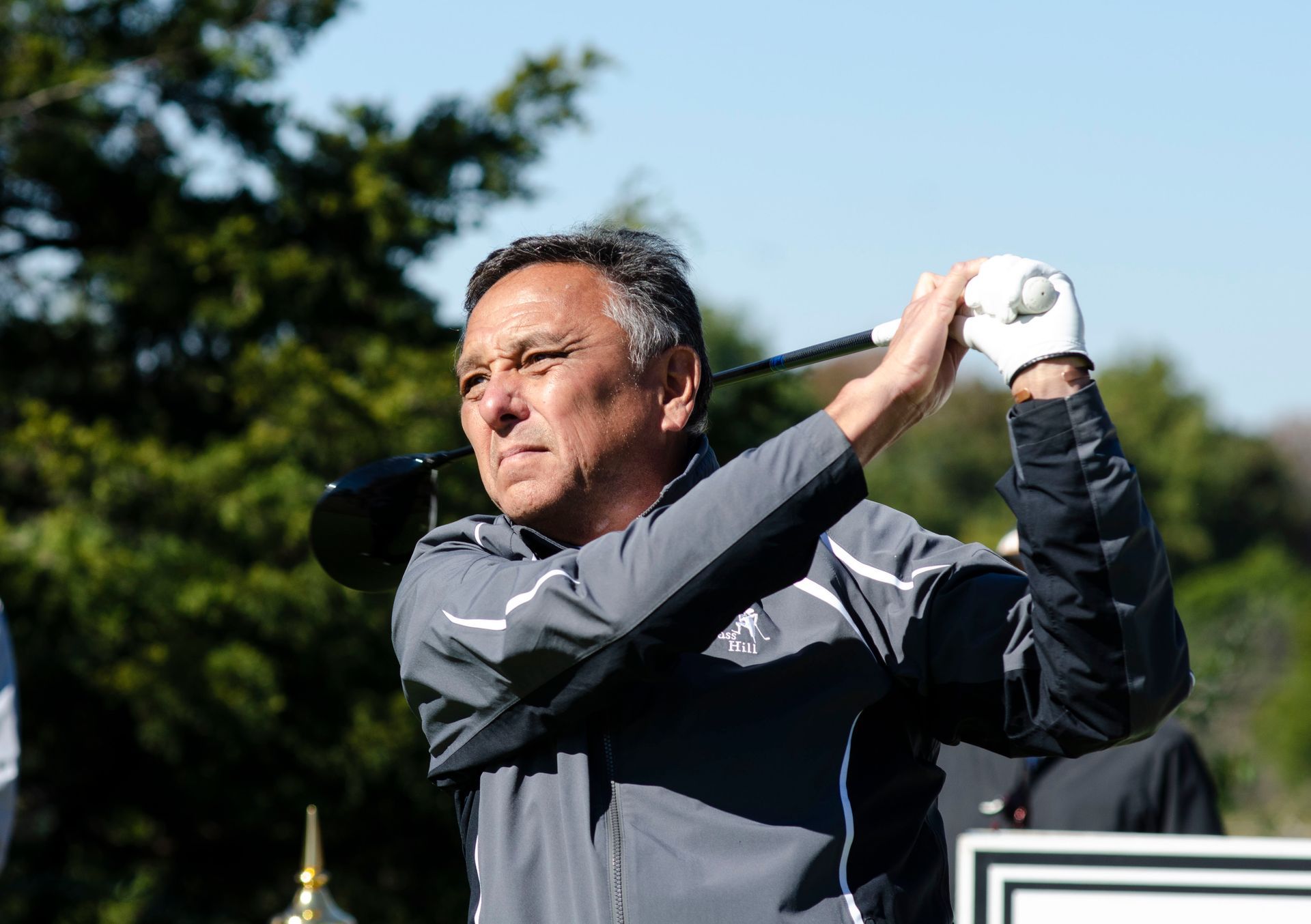 Golfer in dark jacket swinging a club outdoors against a blue sky.