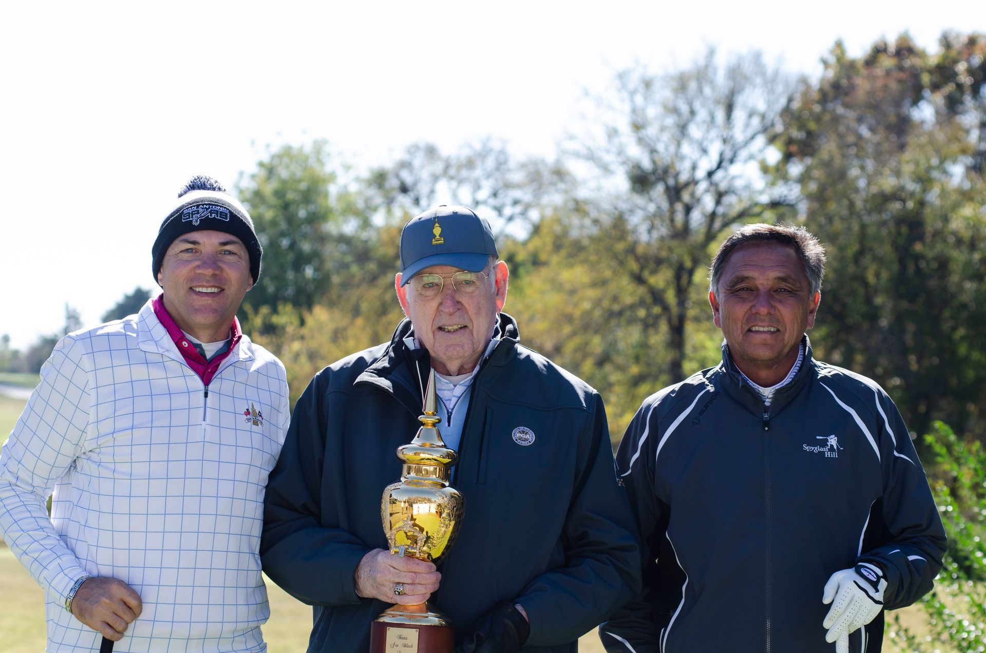 Three people outdoors holding a trophy, with trees in the background.