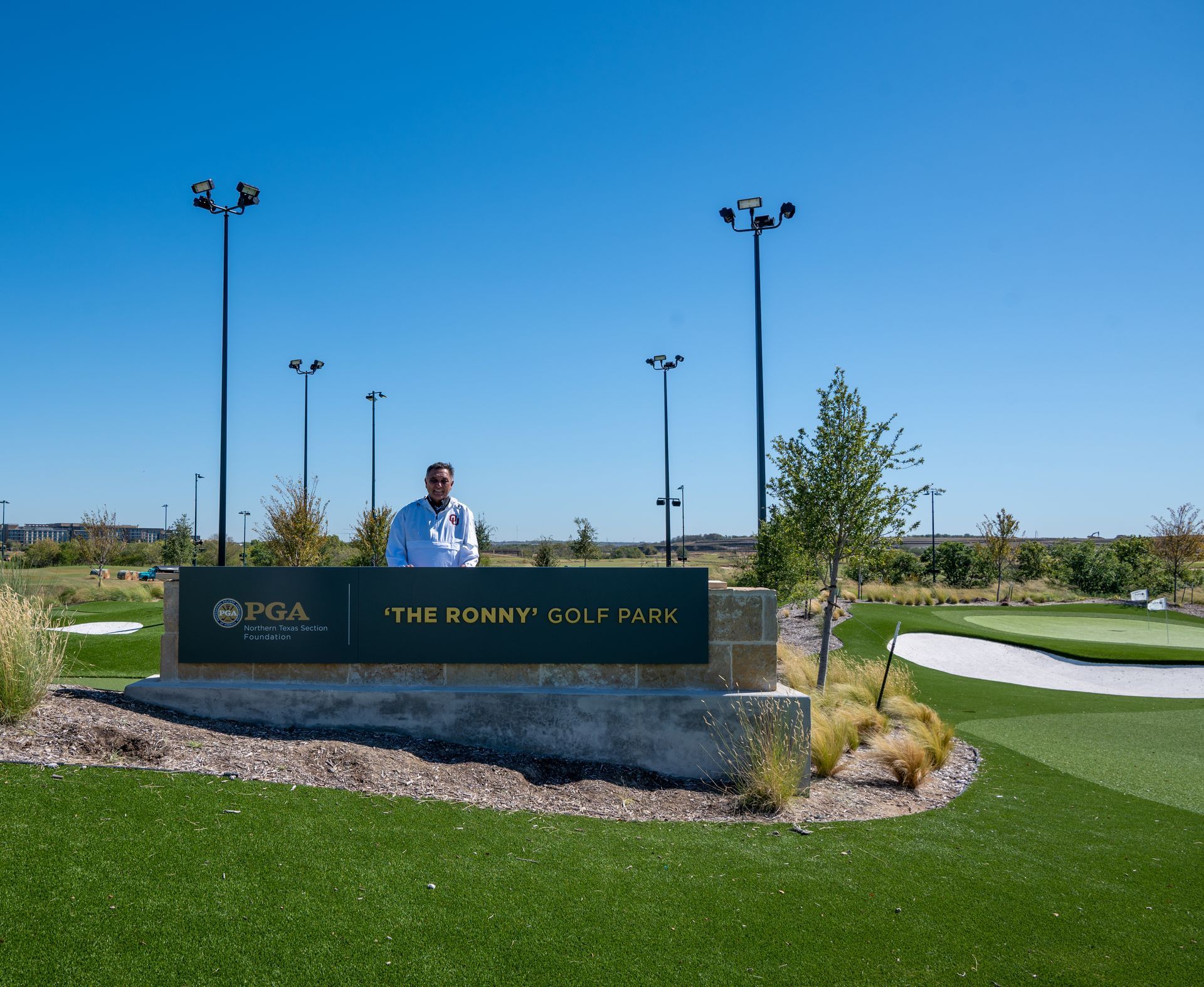 Man standing behind a golf course sign in a sunny landscaped park with green turf and light poles