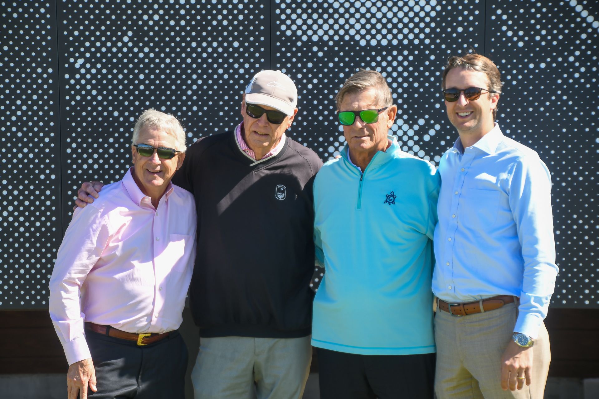 Four people posing outdoors, wearing sunglasses and colorful shirts, standing in front of a patterned wall.
