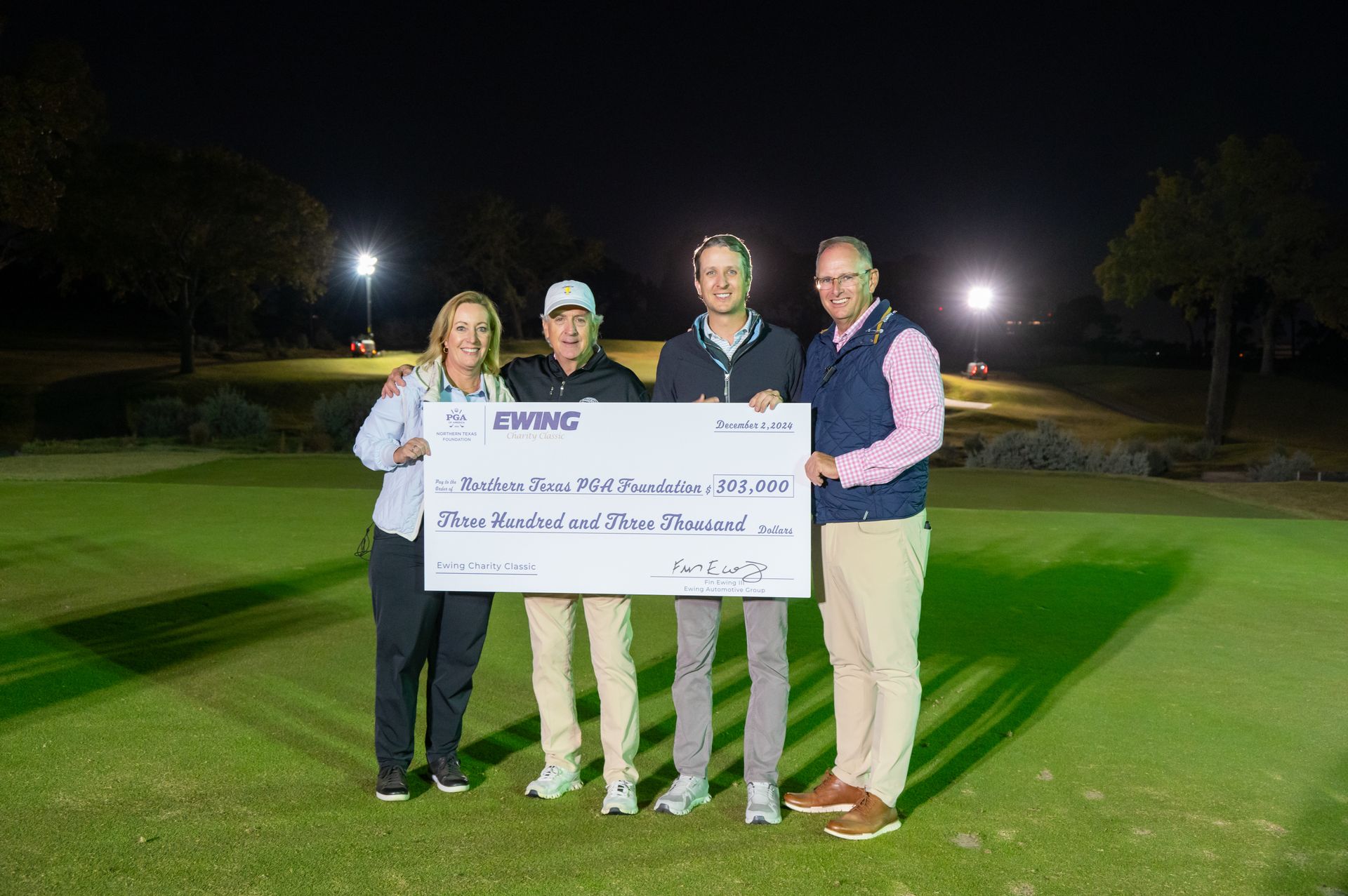 Four people holding a large check on a lit golf course at night