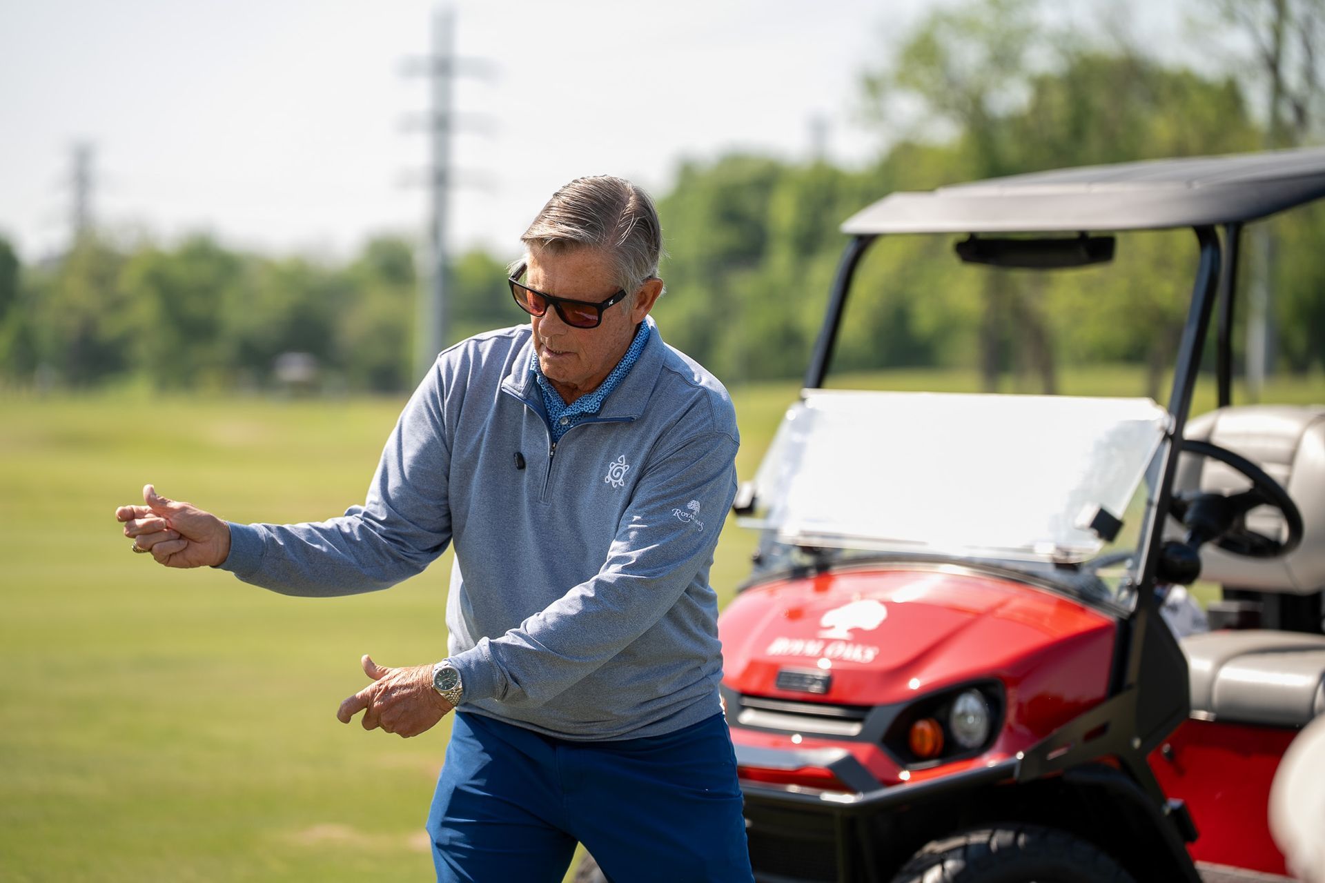 Man in blue shirt swinging a golf club beside a red golf cart on a grassy course