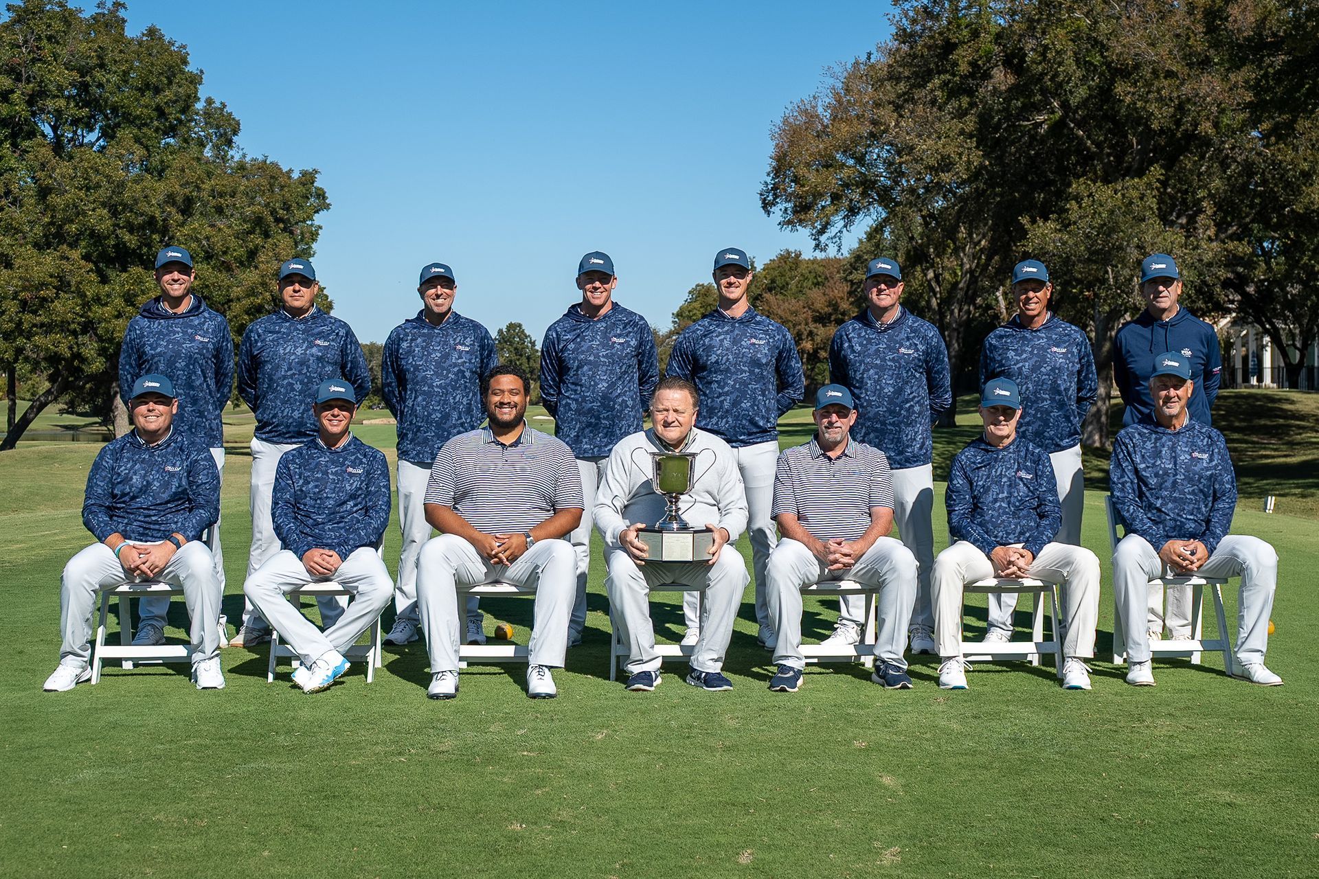 Cricket team posing outdoors on a grassy field in blue uniforms and white pants.
