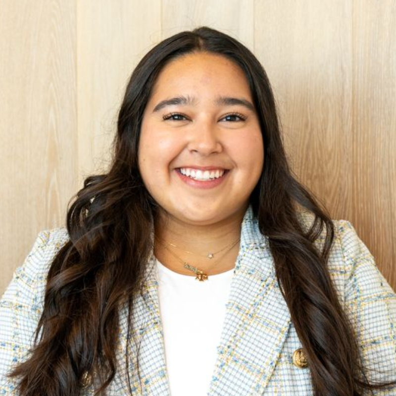 Smiling woman with long dark hair wearing a plaid blazer against a light wood background