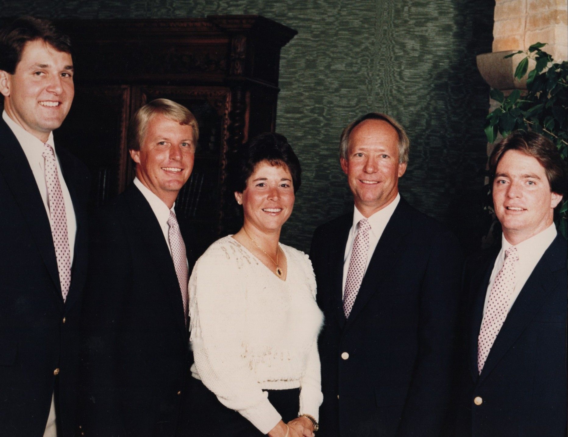 Five people in formal attire posing indoors, with a green patterned wall and a wooden cabinet behind them.