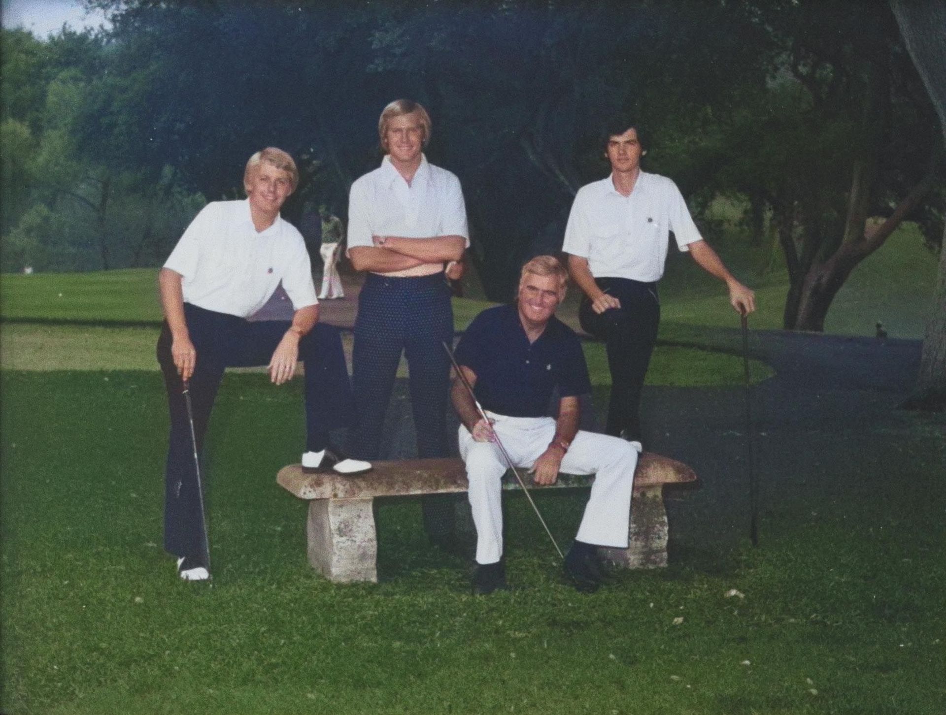 Four people in white and dark shirts posing around a stone bench on a grassy lawn at dusk.