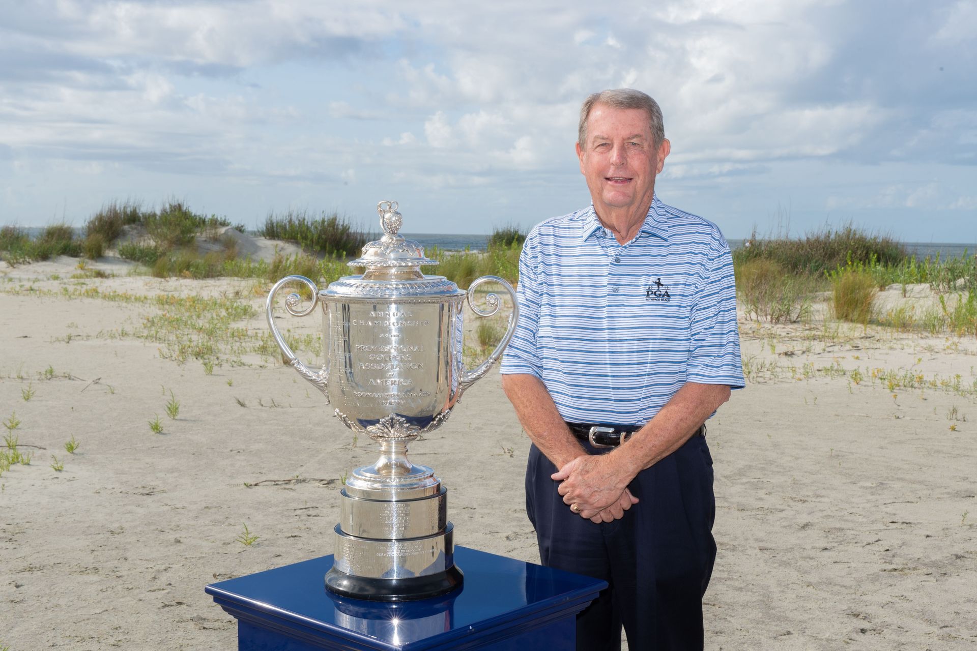 Man standing beside a large silver trophy on a blue pedestal at a sandy beach
