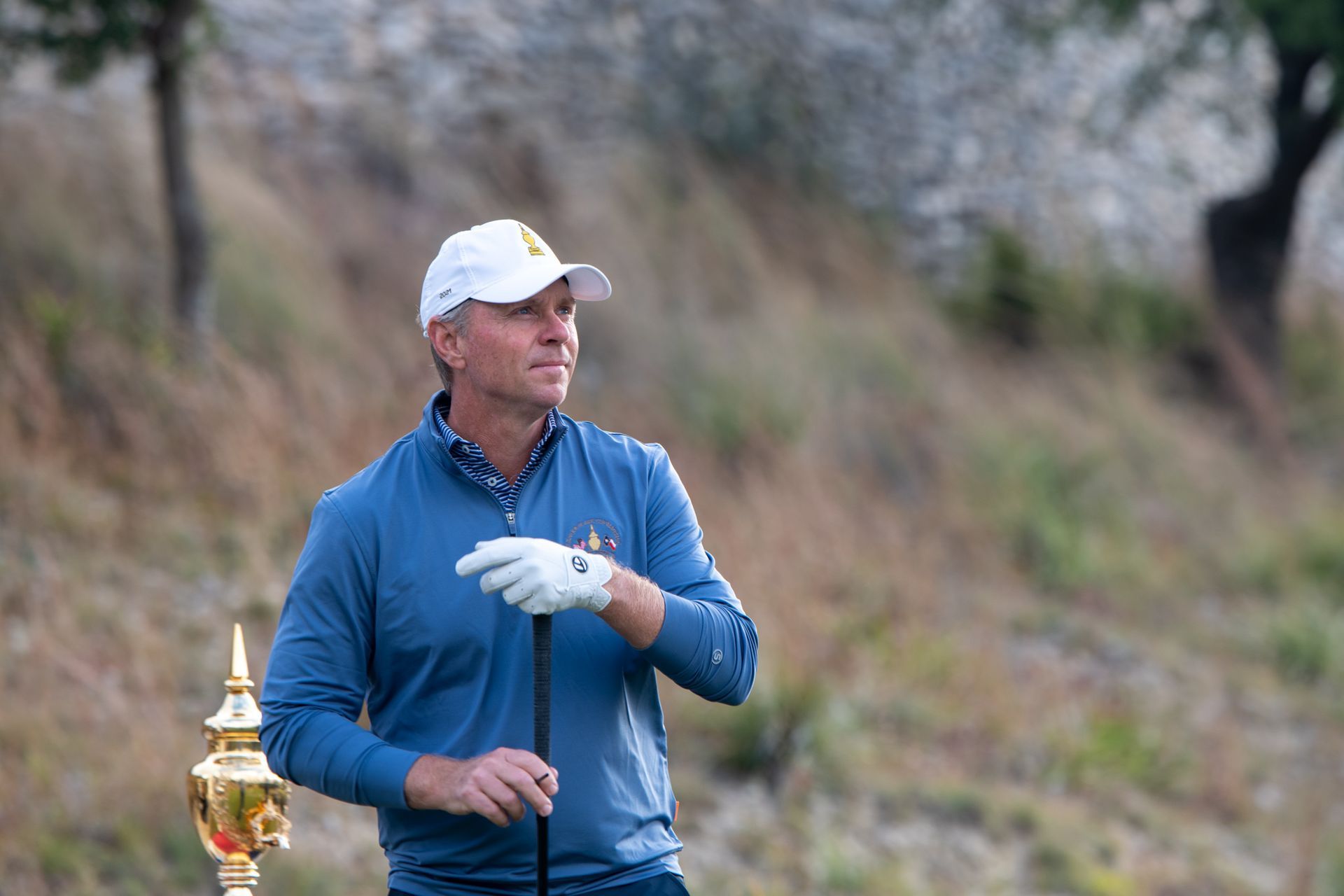 Golfer in blue jacket and white cap holding a club, with a gold trophy beside him on a grassy course.