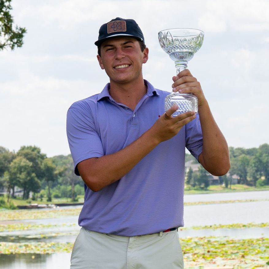 Golfer holding a clear trophy on a course, wearing a purple shirt and cap, with water and trees behind him