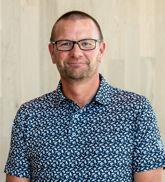 Man wearing glasses and a blue patterned shirt, standing against a light wood wall and smiling at the camera