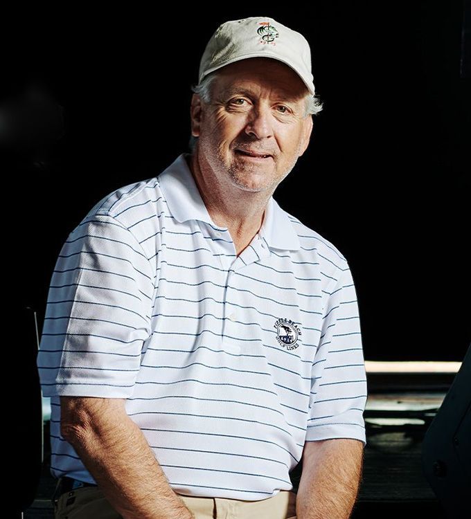 Man in a white striped polo and cap, seated in a dim setting, facing the camera and smiling.