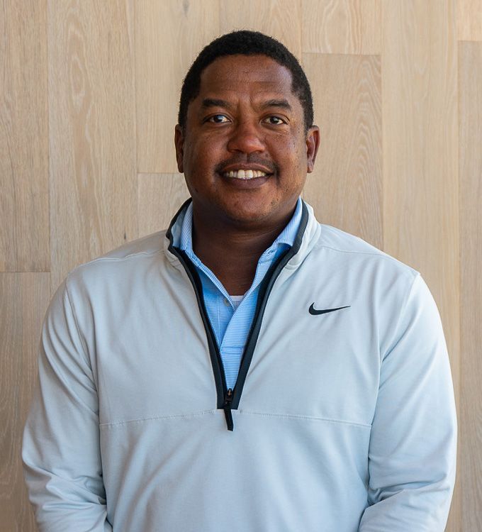 Smiling person in a light gray Nike jacket standing against a wood-paneled wall