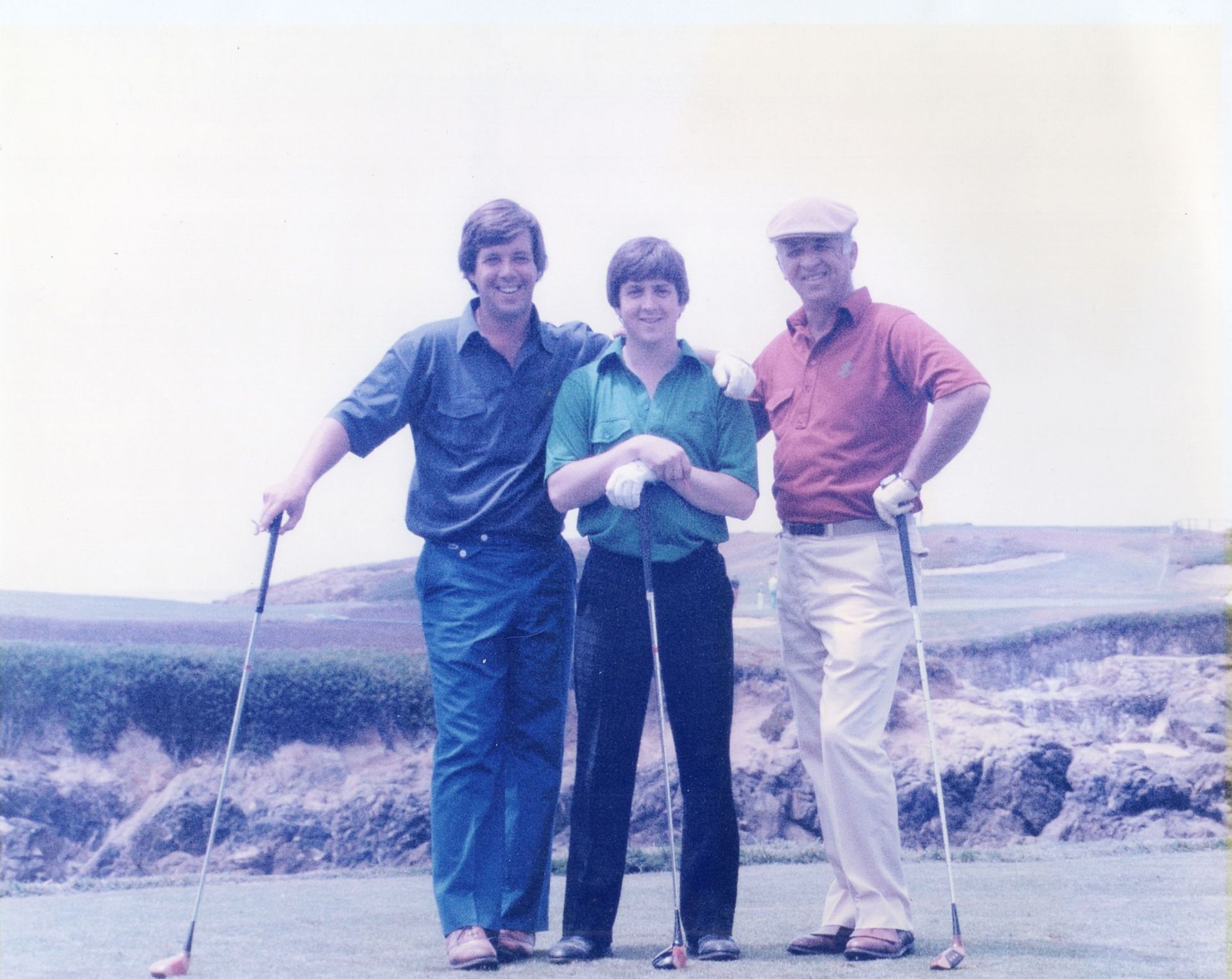 Three people standing together outdoors on a golf course, holding clubs and smiling.