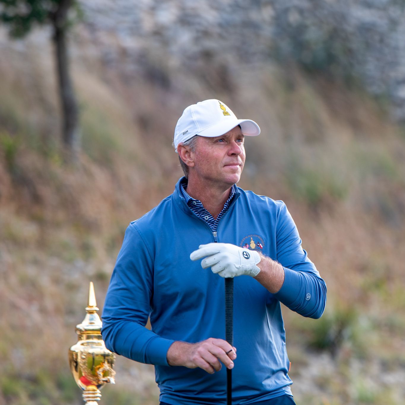 Golfer in blue jacket and white cap holding a club, with a gold trophy beside him on a grassy course.