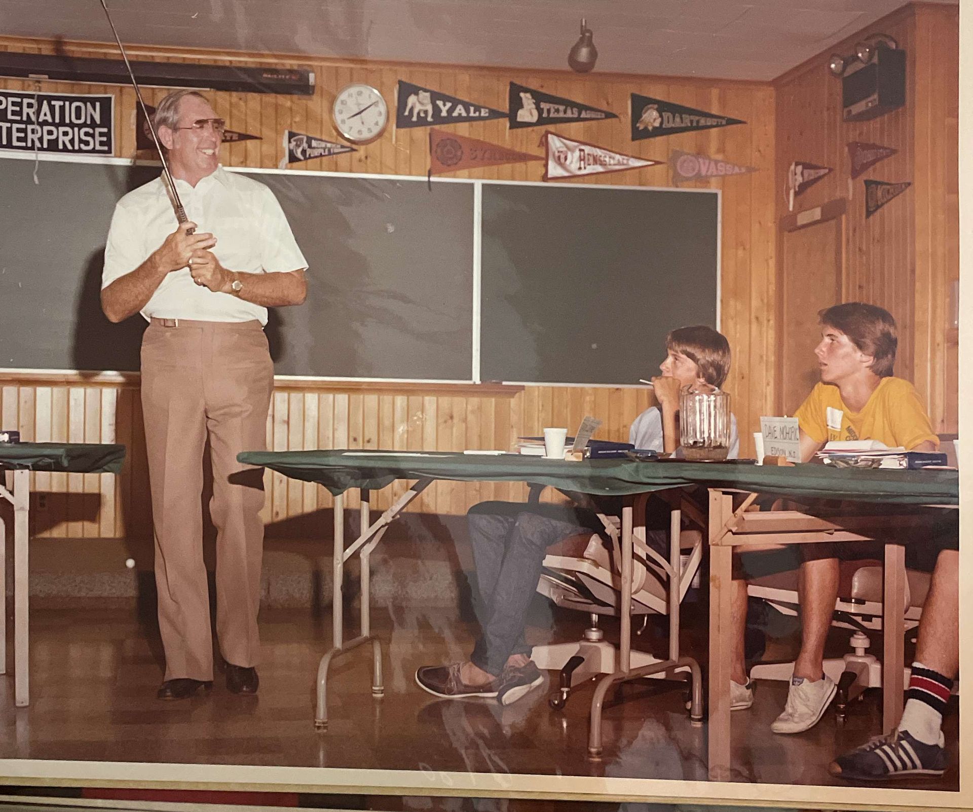 Teacher standing in a classroom beside a chalkboard while students sit at desks and listen.