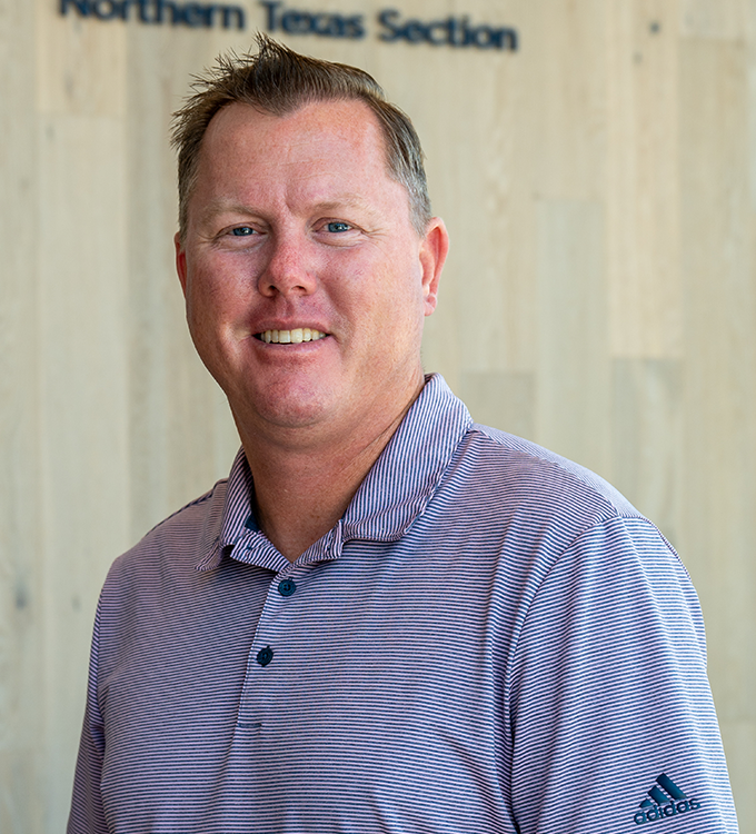 Smiling man in a striped polo standing indoors with a wood wall backdrop.