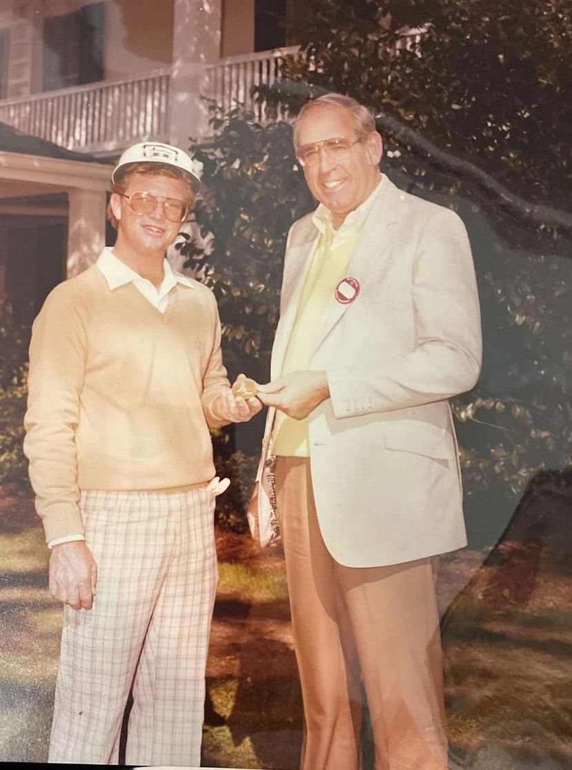 Two men shaking hands outdoors at night, one in a beige suit and the other in a sweater and plaid pants.