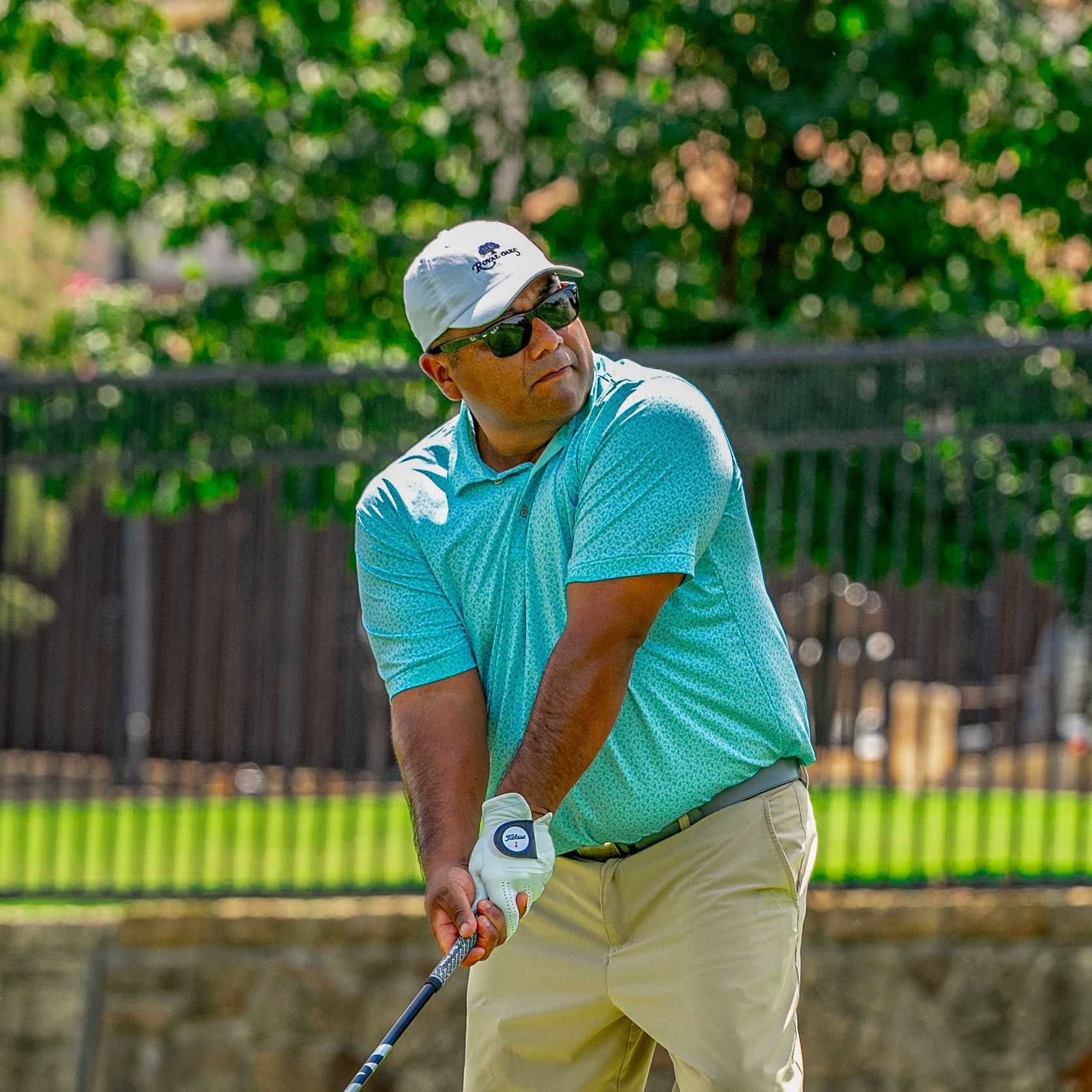 Golfer mid-swing on a course, wearing a blue shirt and white cap, with driver extended against a cloudy sky